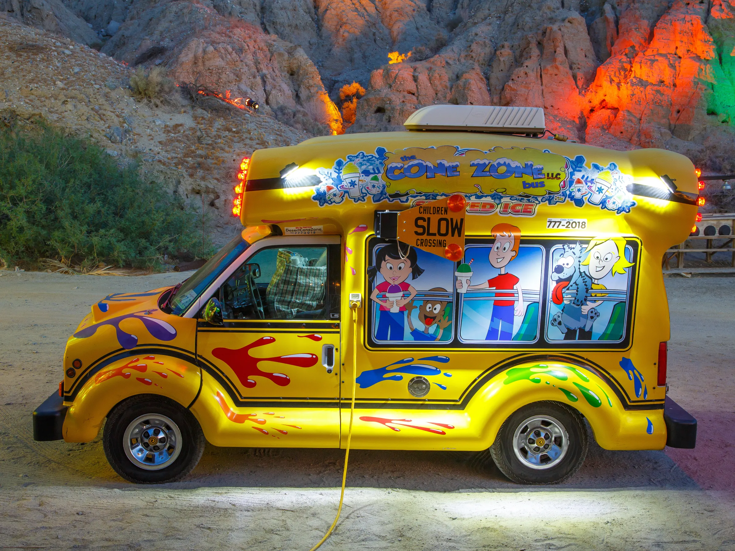 Bright yellow ice cream truck with colorful graphics parked in illuminated rocky area.