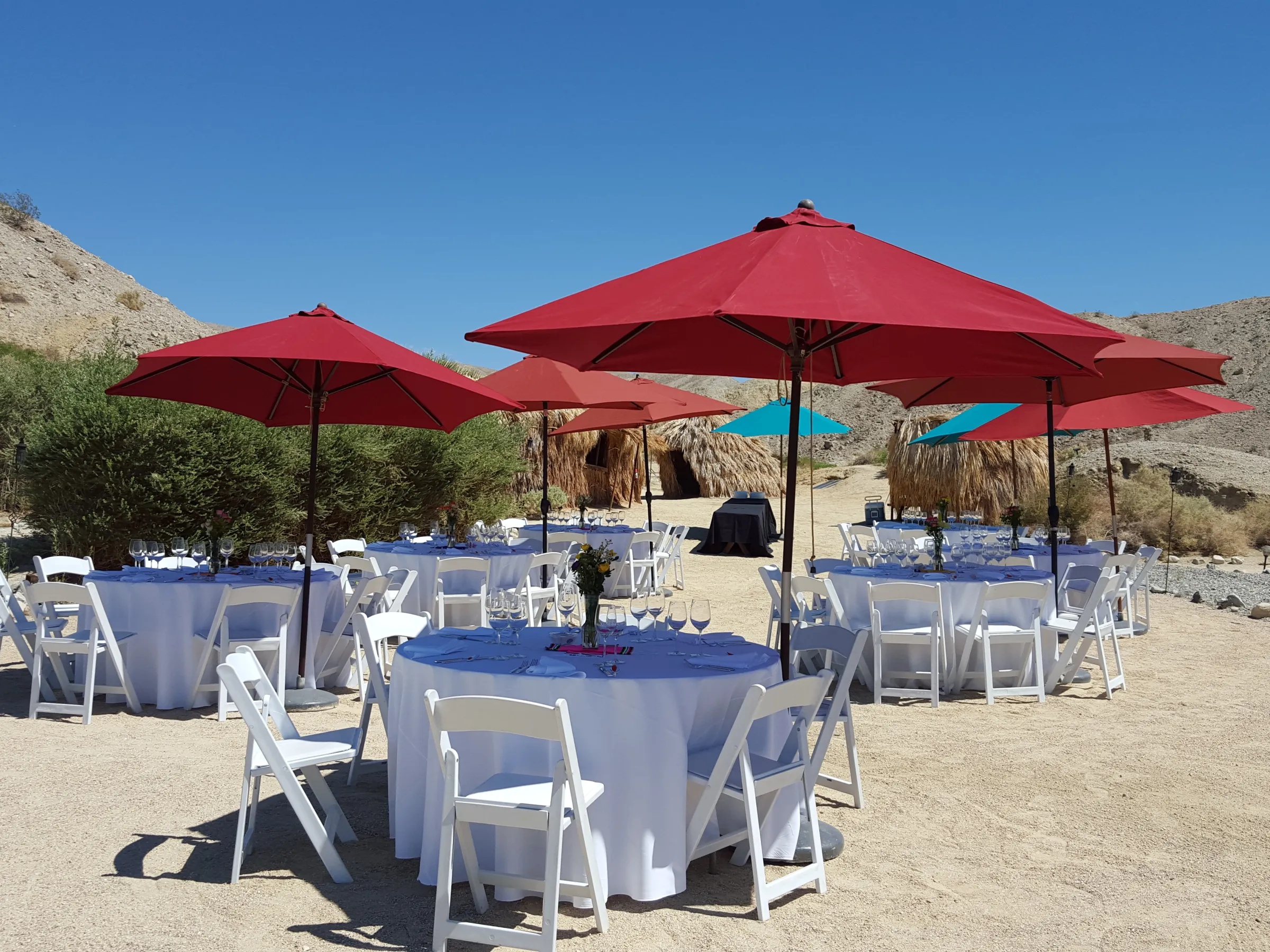 Outdoor desert setup with round tables, red umbrellas, and white chairs.