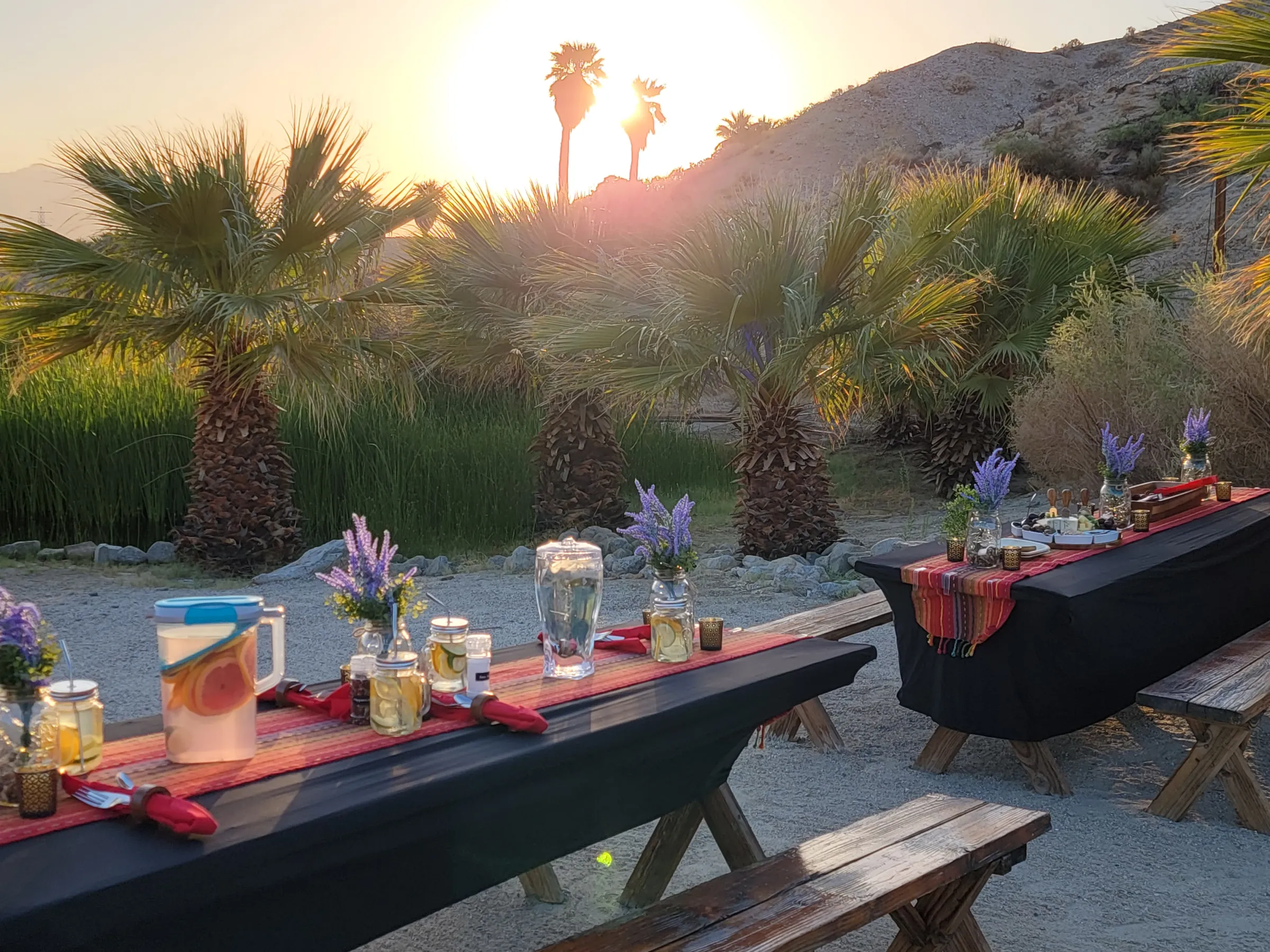Outdoor dining setup with tables, decorations, and sunset over palm trees and desert.