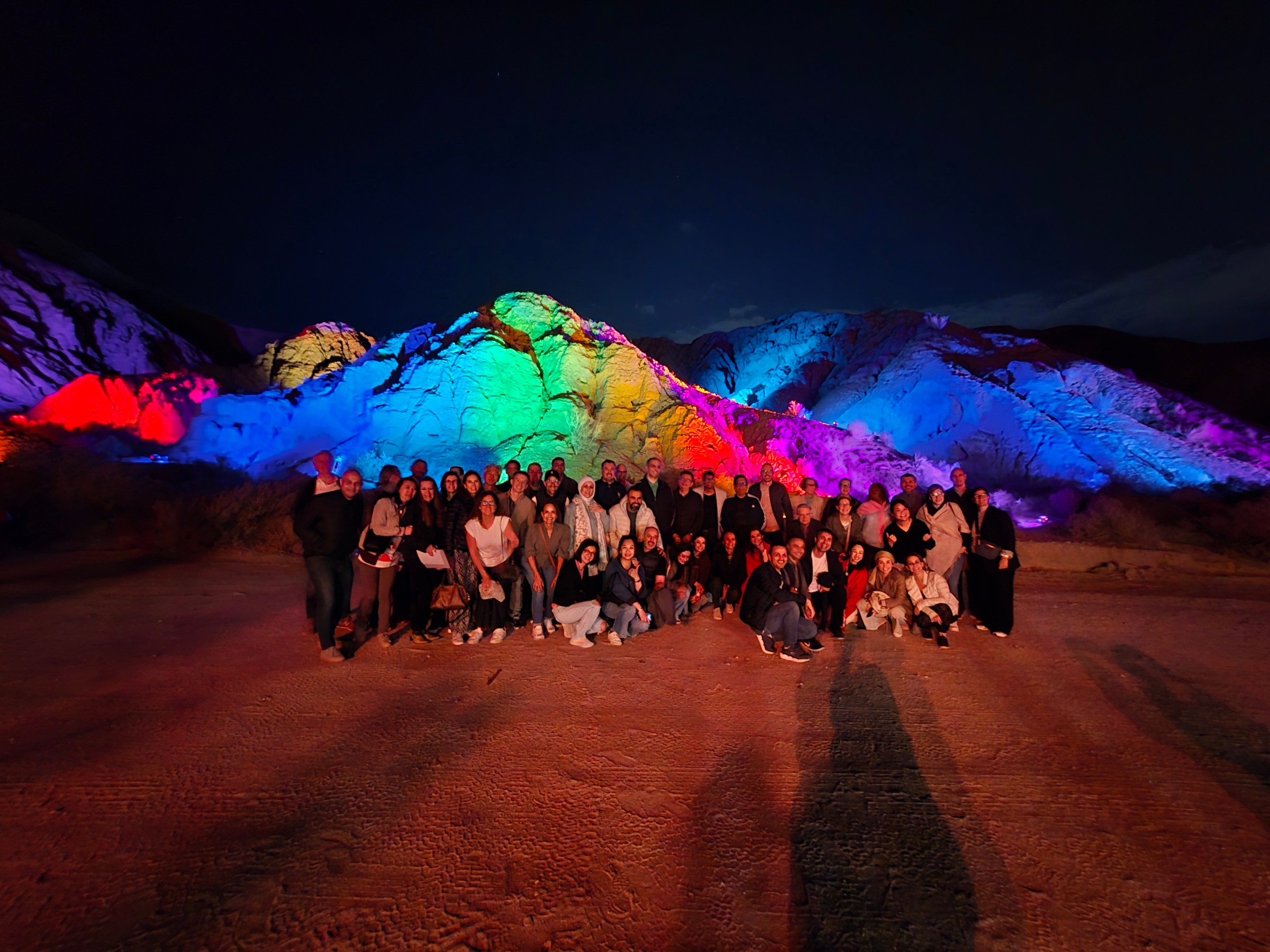 Group of people posing at night with colorful lit mountains in the background.