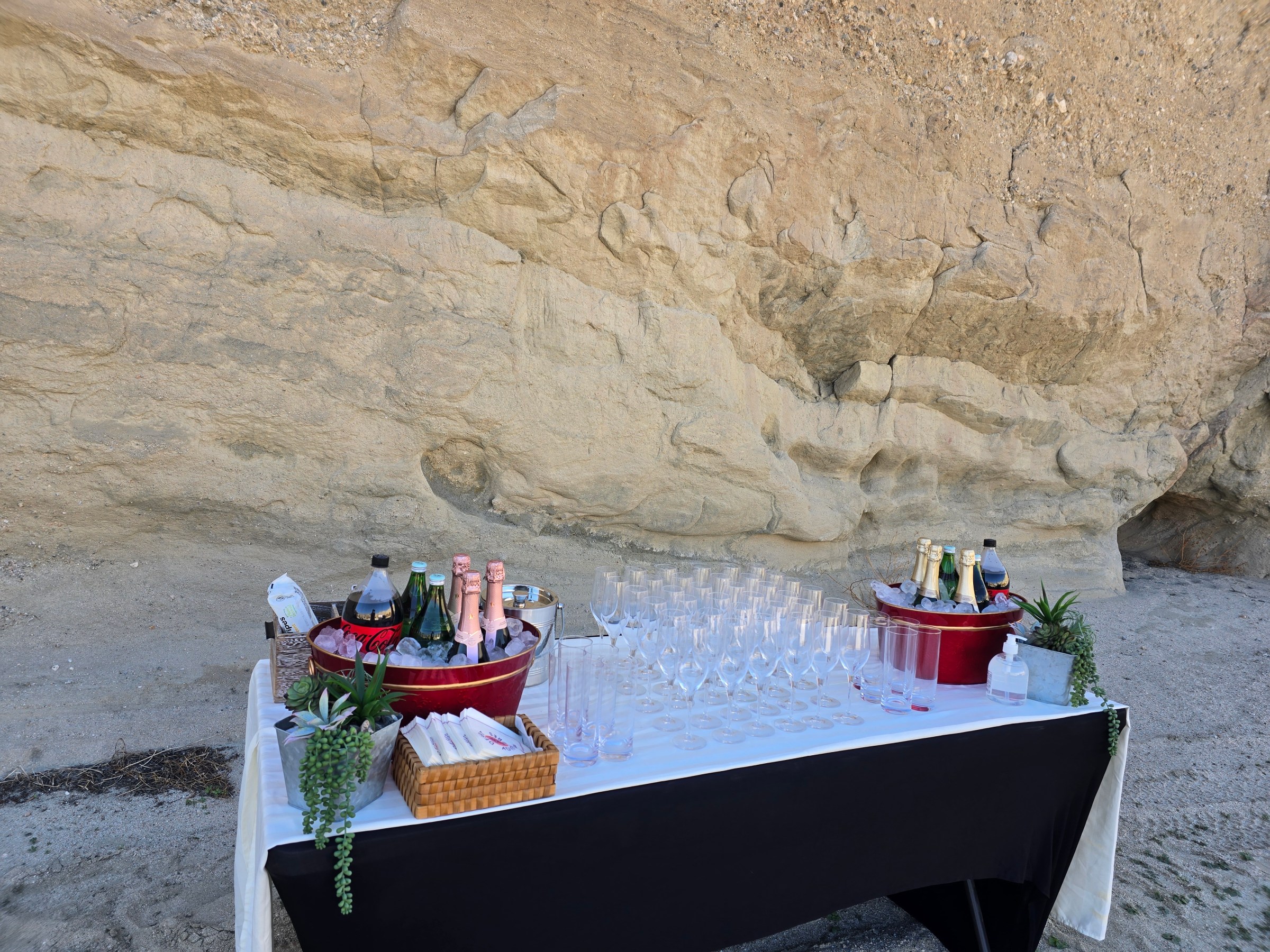 Outdoor refreshment table with drinks, glasses, and decorations against a rock wall.