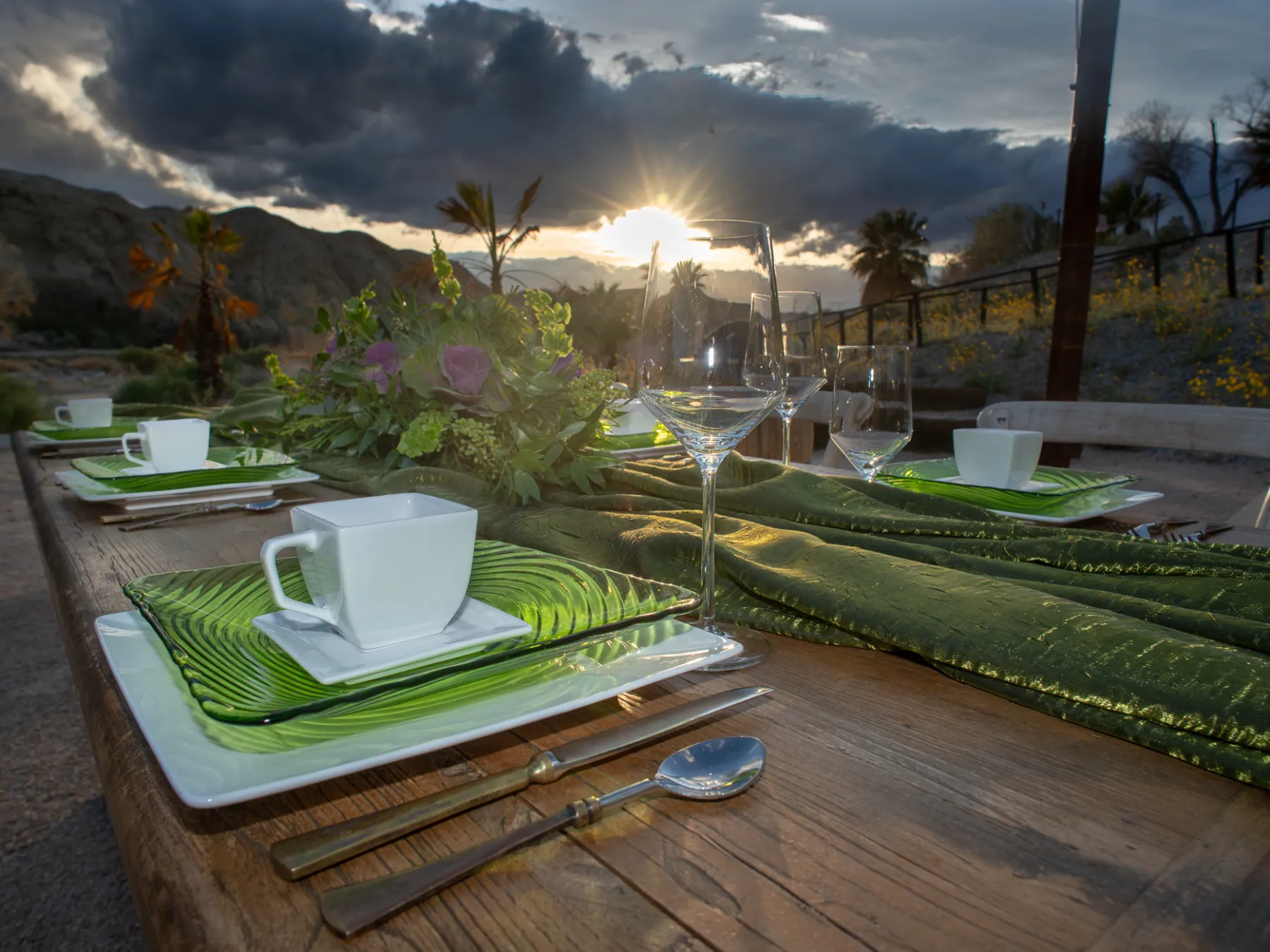 Outdoor table setting with cups and plates, green accents, and sunset view.