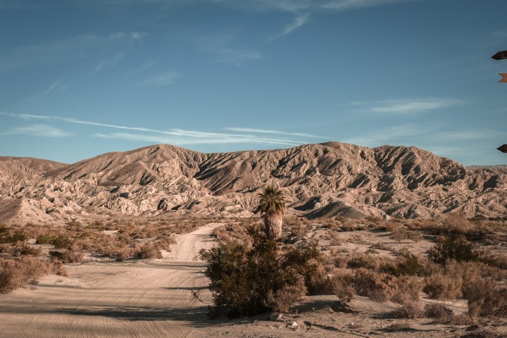 Desert landscape with rugged mountains, sandy terrain, and scattered vegetation under a clear blue sky.