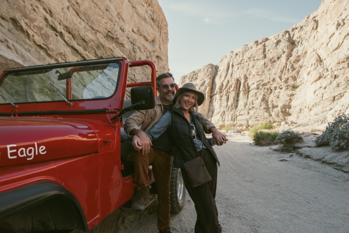 Couple smiling beside red jeep in rocky canyon.