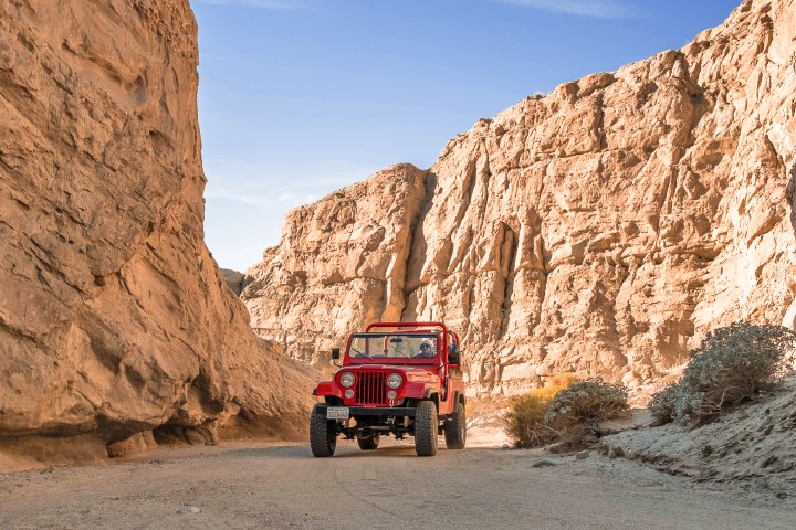 Red off-road vehicle driving through narrow desert canyon with rocky walls.