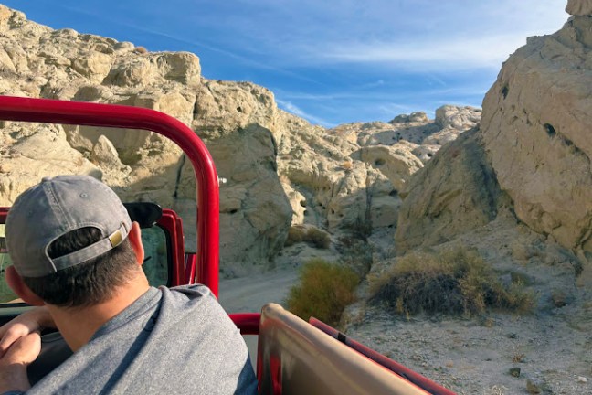 Person in a convertible jeep driving through rocky desert terrain under a clear blue sky.