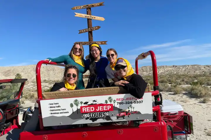 Five people posing on a red jeep with desert landscape and directional sign in the background.